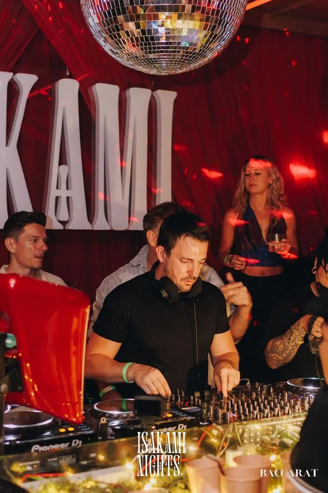 Isakami Nights at Baccarat Bangkok — DJ at the booth with Pioneer CDJs, the giant ISAKAMI letters and disco ball behind, red curtains and Baccarat venue branding